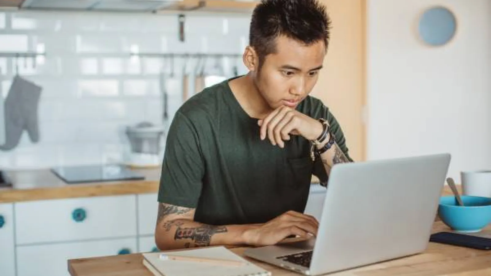 A young man with spiky black hair and tatoos wearing a dark green t-shirt works at a laptop