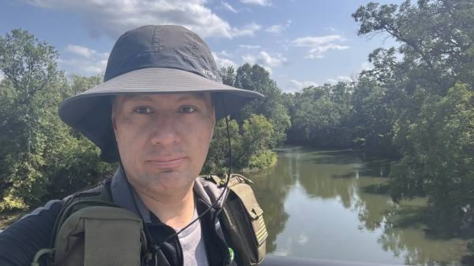 A man wearing a black bucket hat and carrying a backpack stands in front of a bridge with green trees and a blue sky with clouds in the background