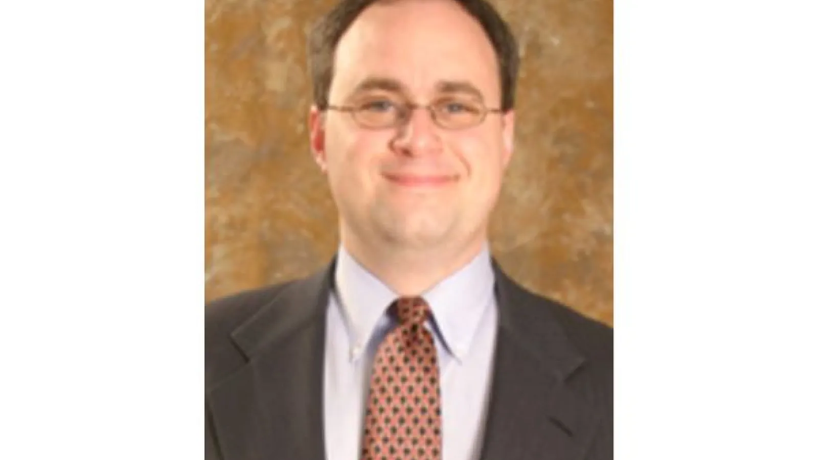 A portrait of Daniel Collins, wearing wire-rim glasses, a suit and red-and-black patterned tie