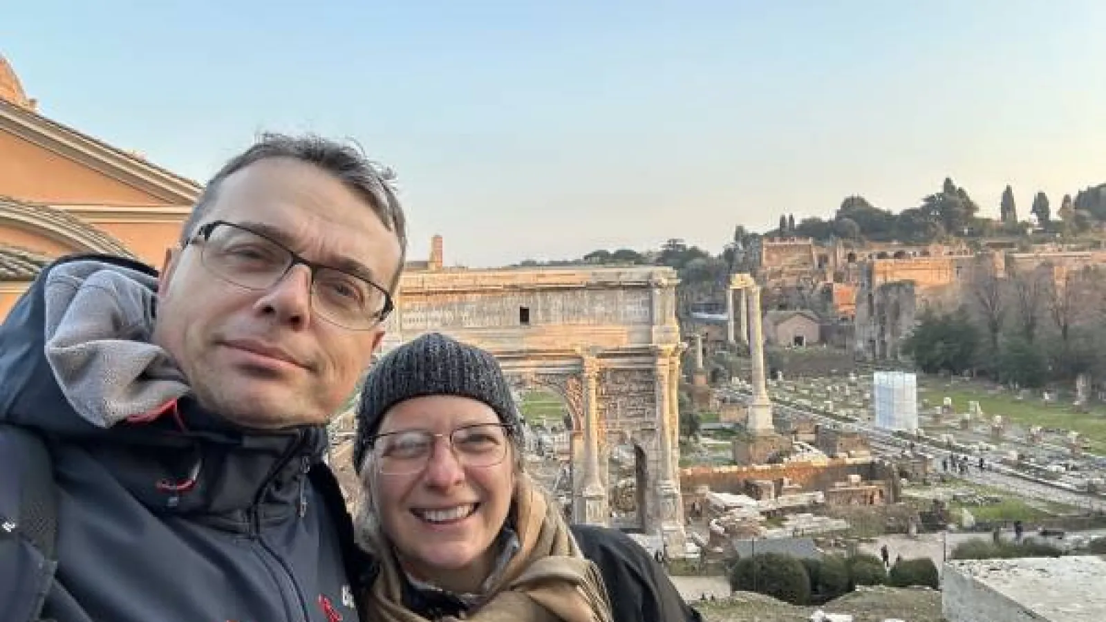 A man and a woman in winter gear stand in front of a tourist site of ancient ruins