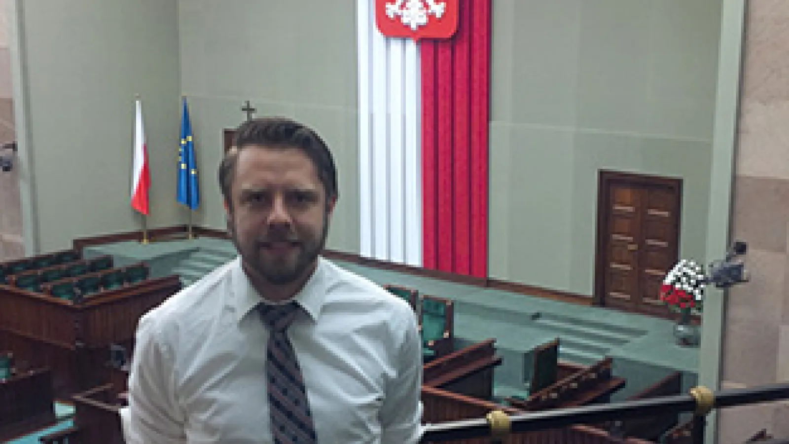 Man standing on a balcony in a large room with the Polish flag hanging in background
