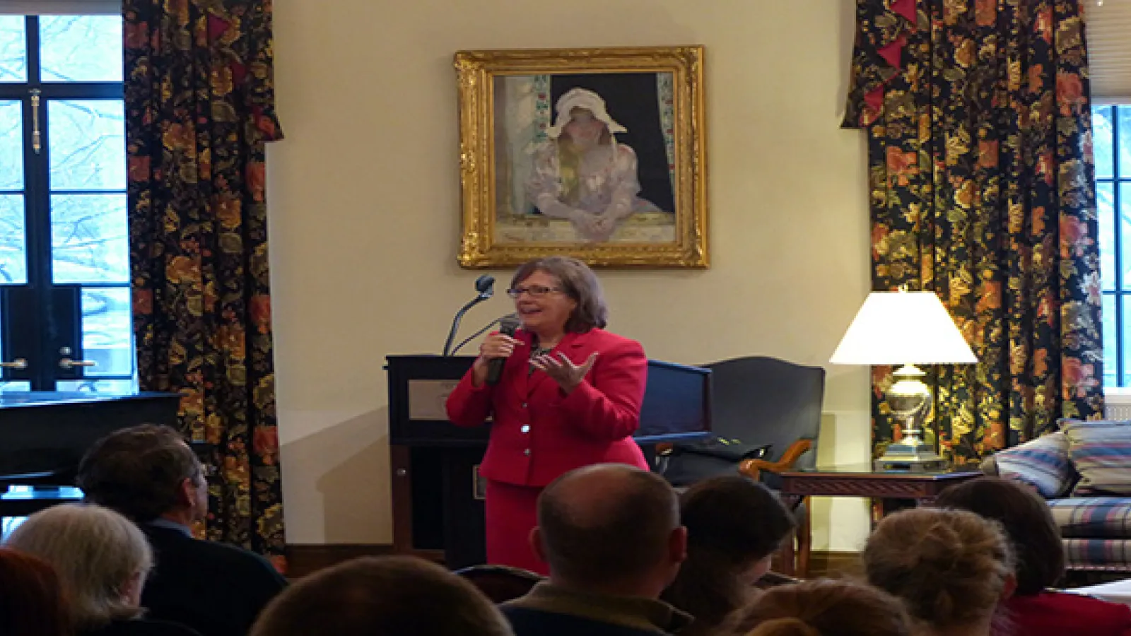 Woman wearing red suit holding microphone and talking in front of a seated audience