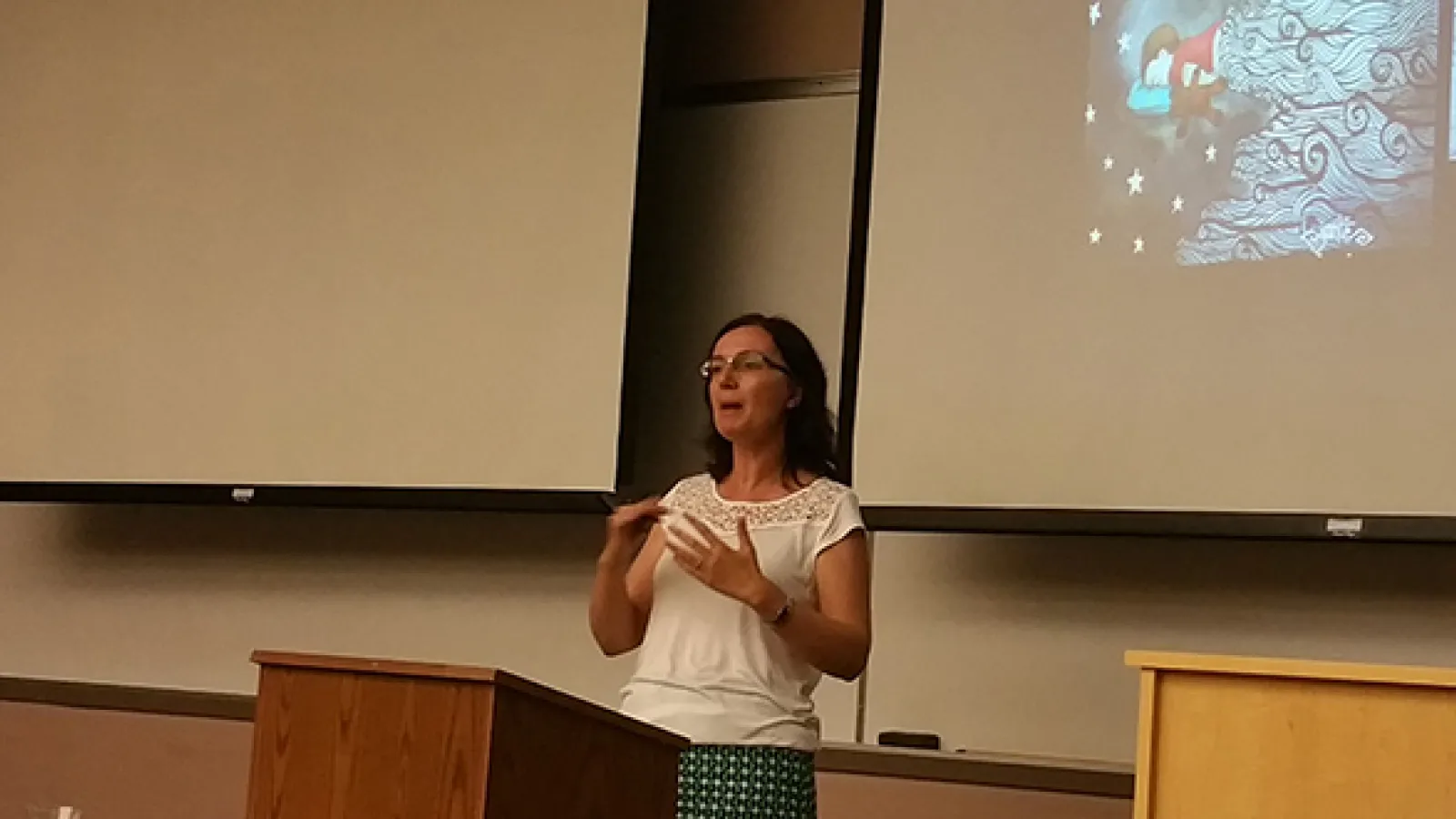 Woman standing at a podium speaking with large projector screens to either side of her