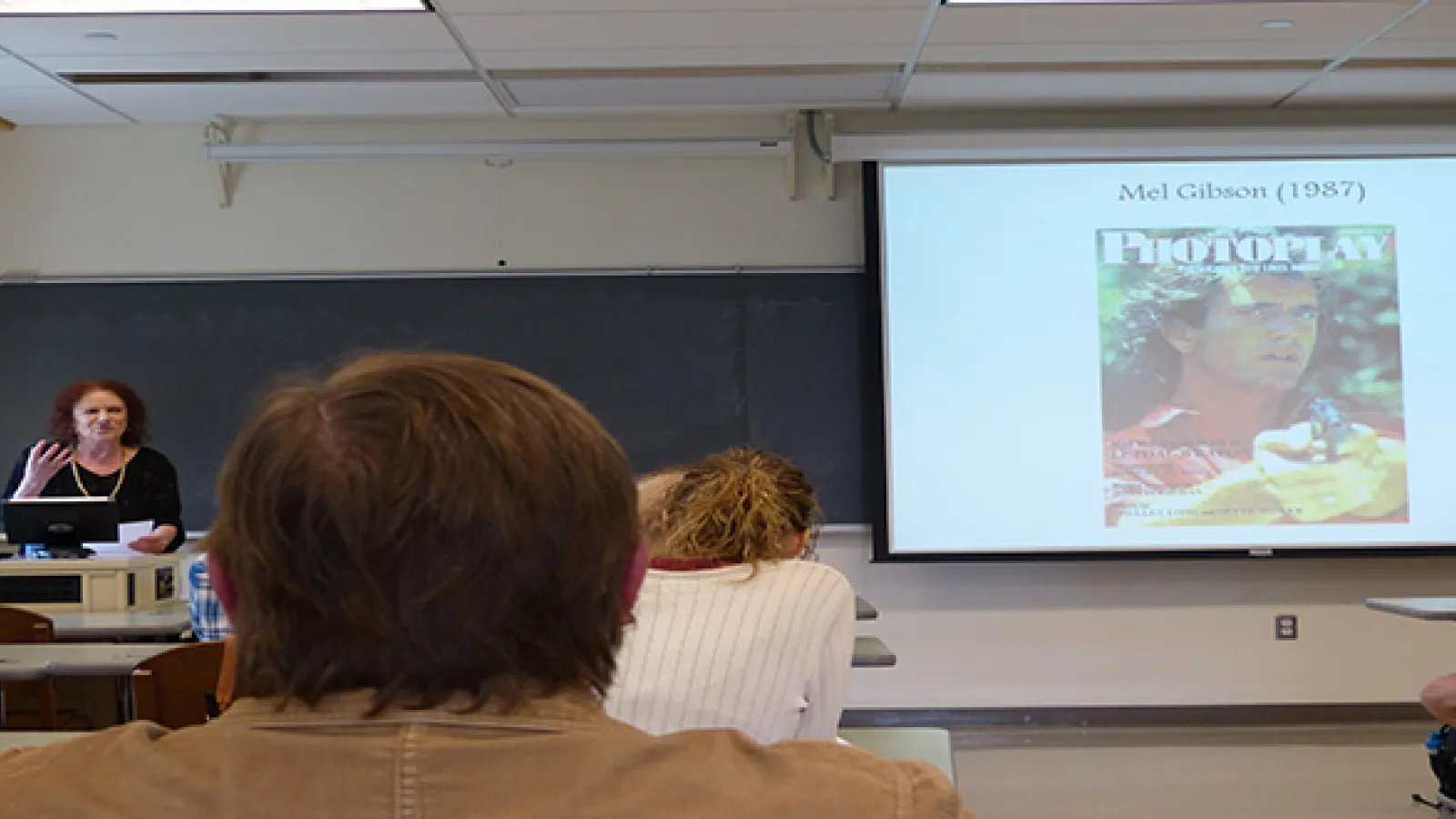 A woman standing at a lecturn speaking with a large image of a film magazine cover showing Mel Gibson at the front of the room