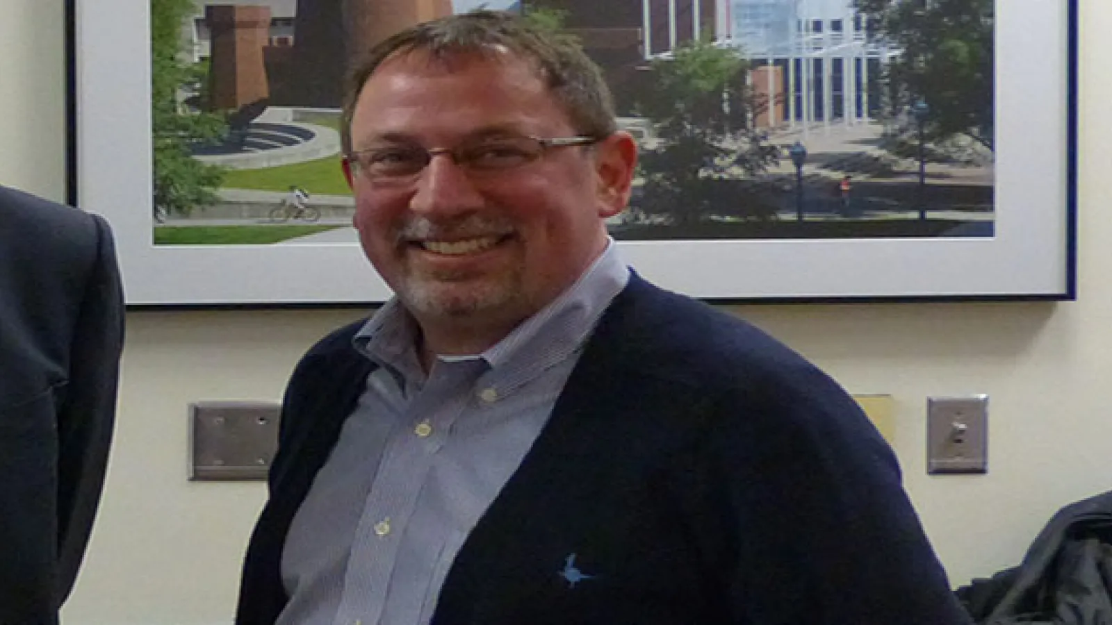 Photo of man's face and torso smiling and wearing glasses with a large picture in the background