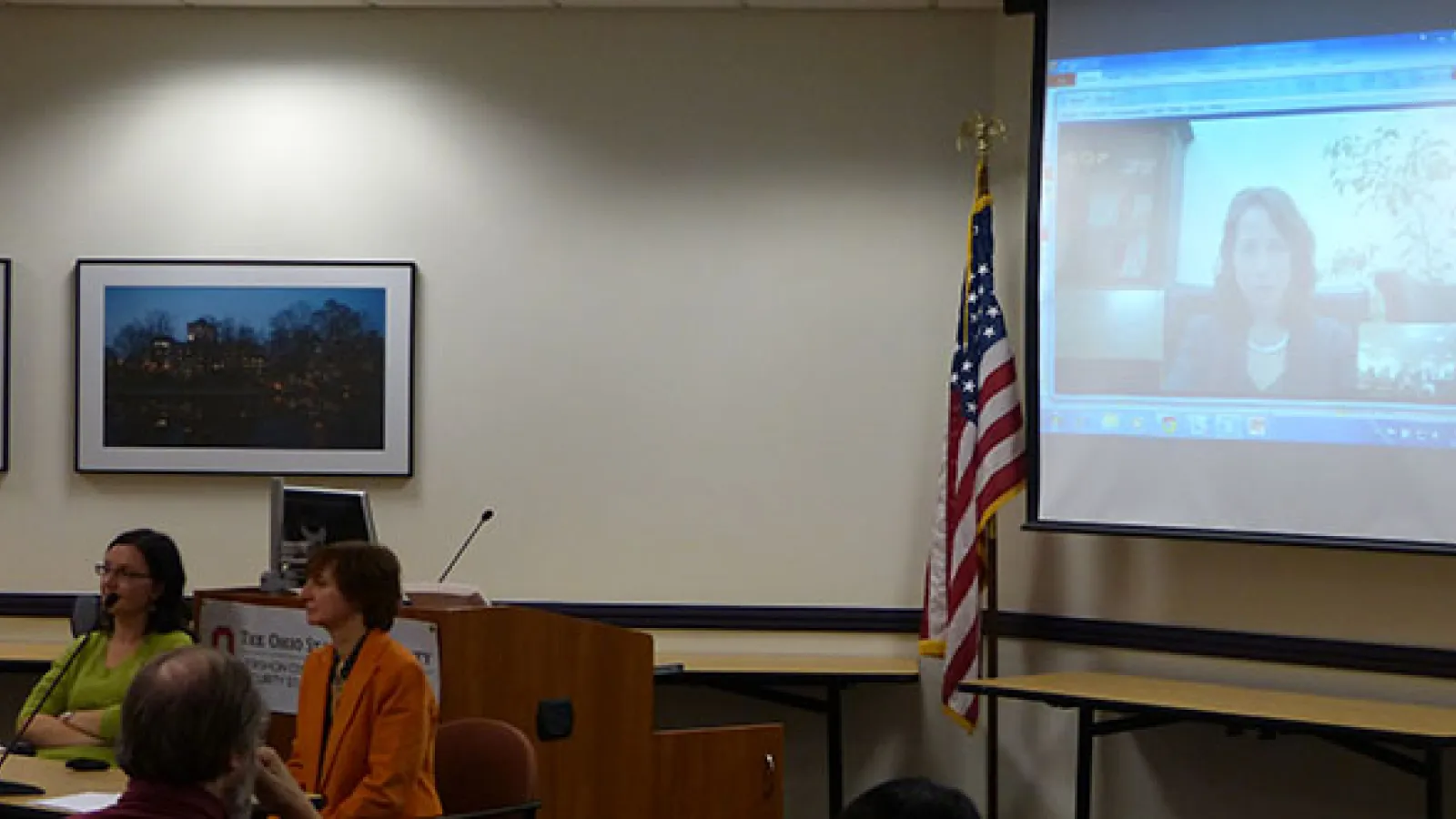 Two women sitting at a table on the left with a large projector screen on the right showing a woman's face speaking
