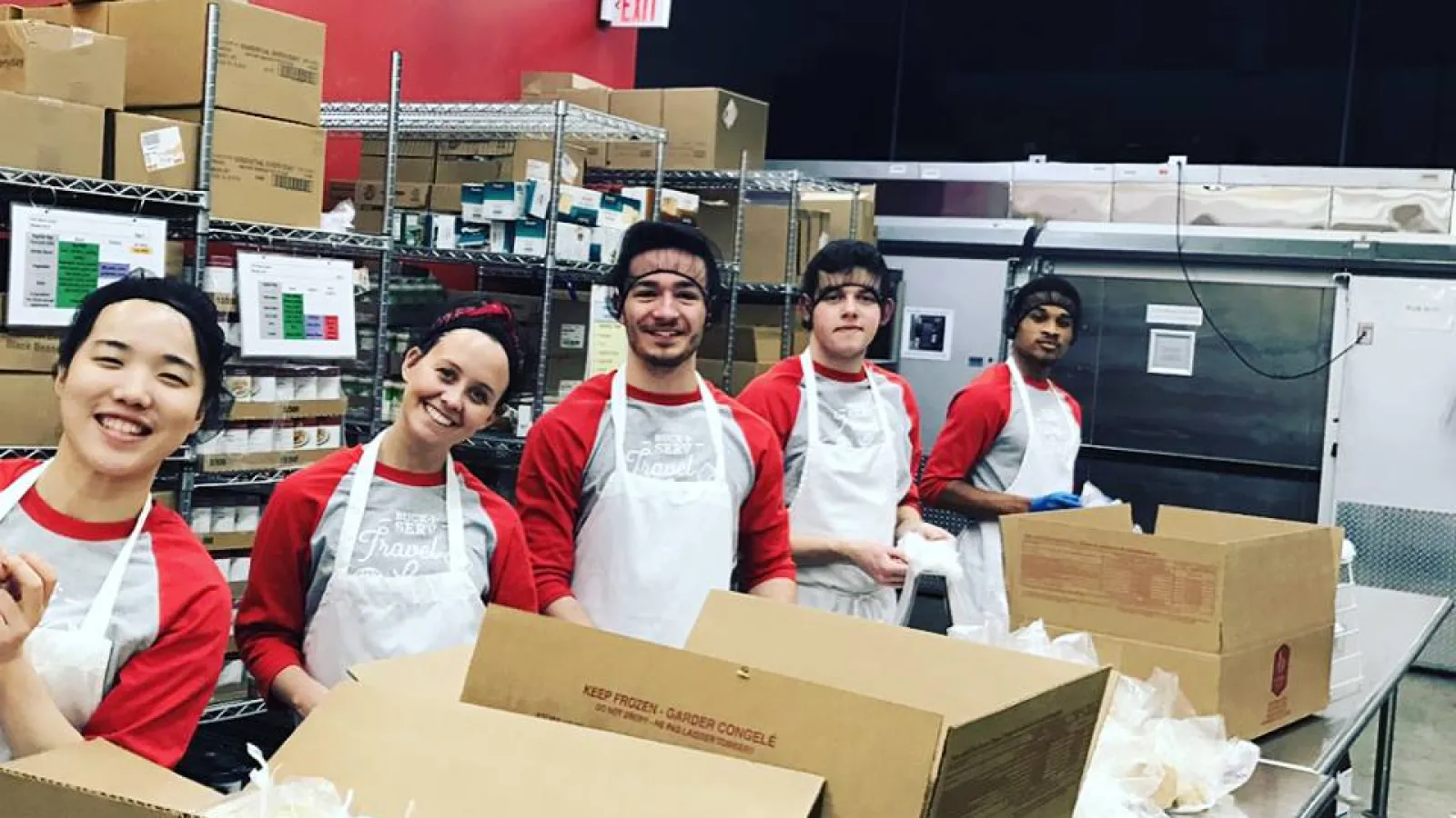5 students packing food at a large table with cardboard boxes