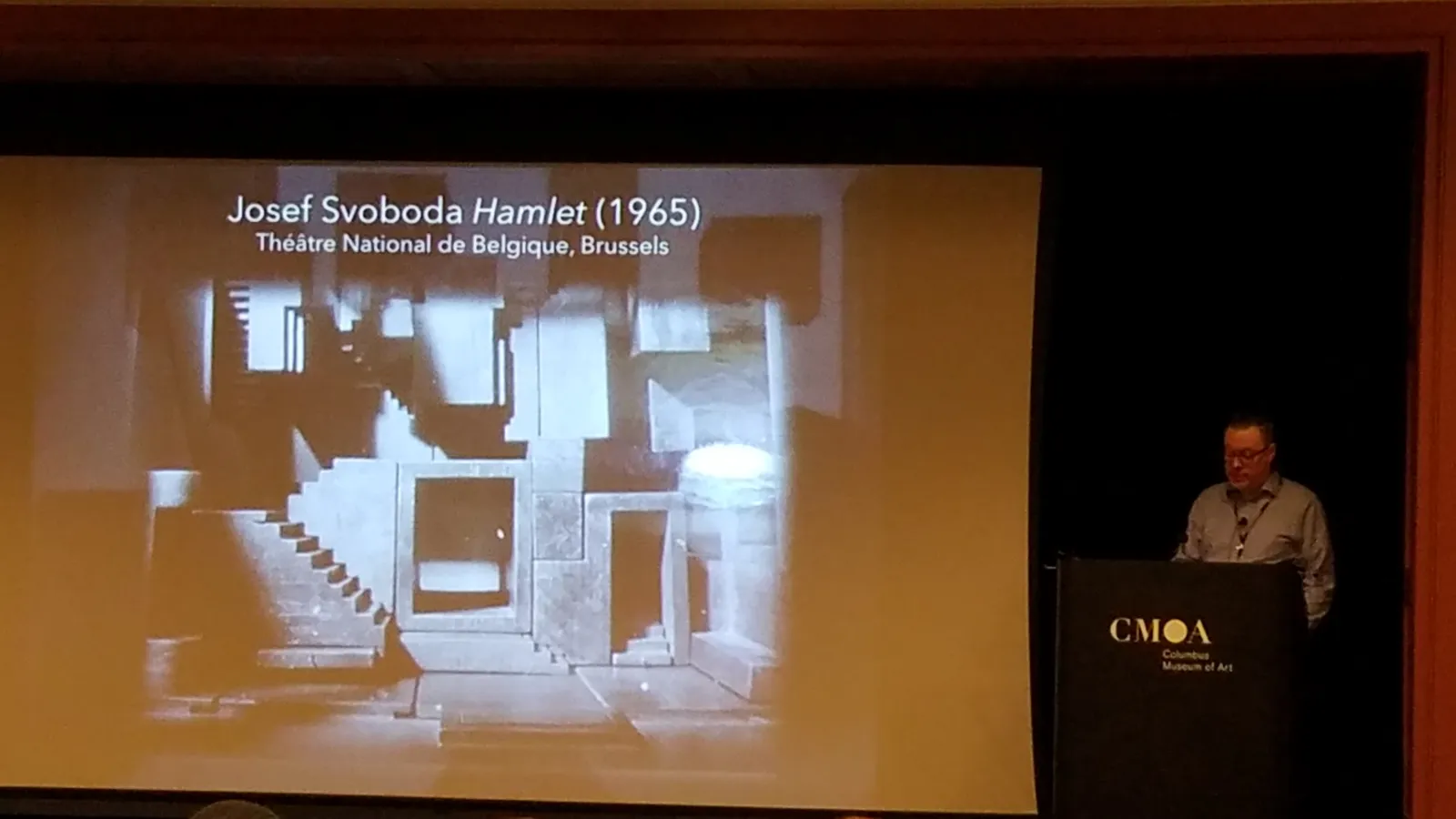 Man standing at a podium talking, to the right of a large projected picture with the words "Josef Svoboda Hamlet 1965"