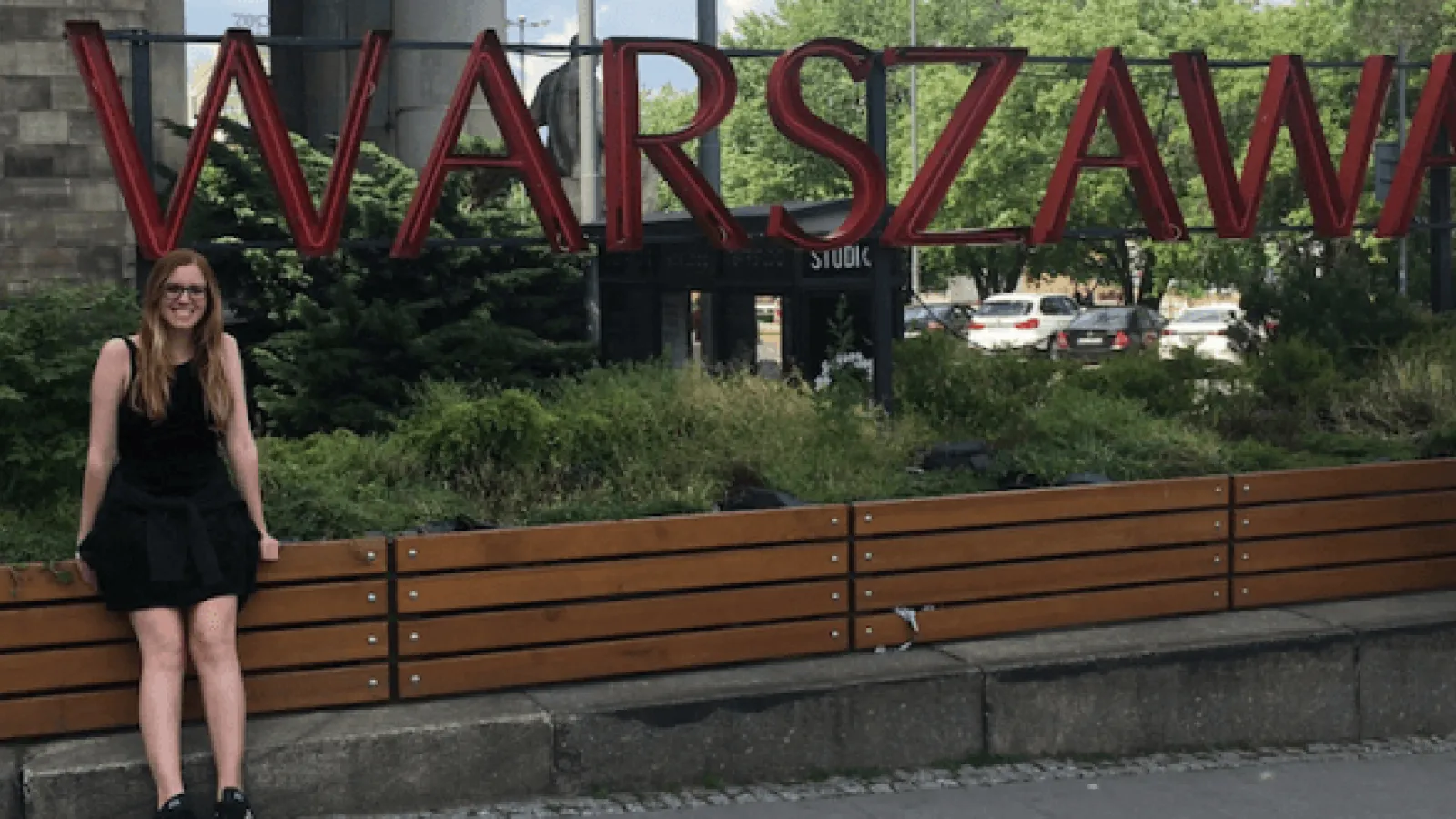 A girl with long hair and glasses standing by a large sign spelling out Warszawa