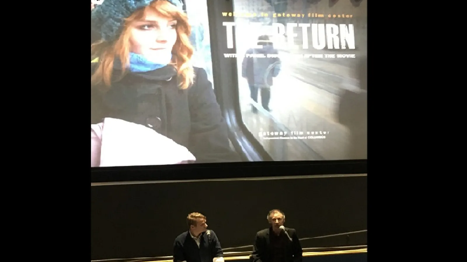2 men sitting on tall stools in front of a large film screen in background showing a girl's face