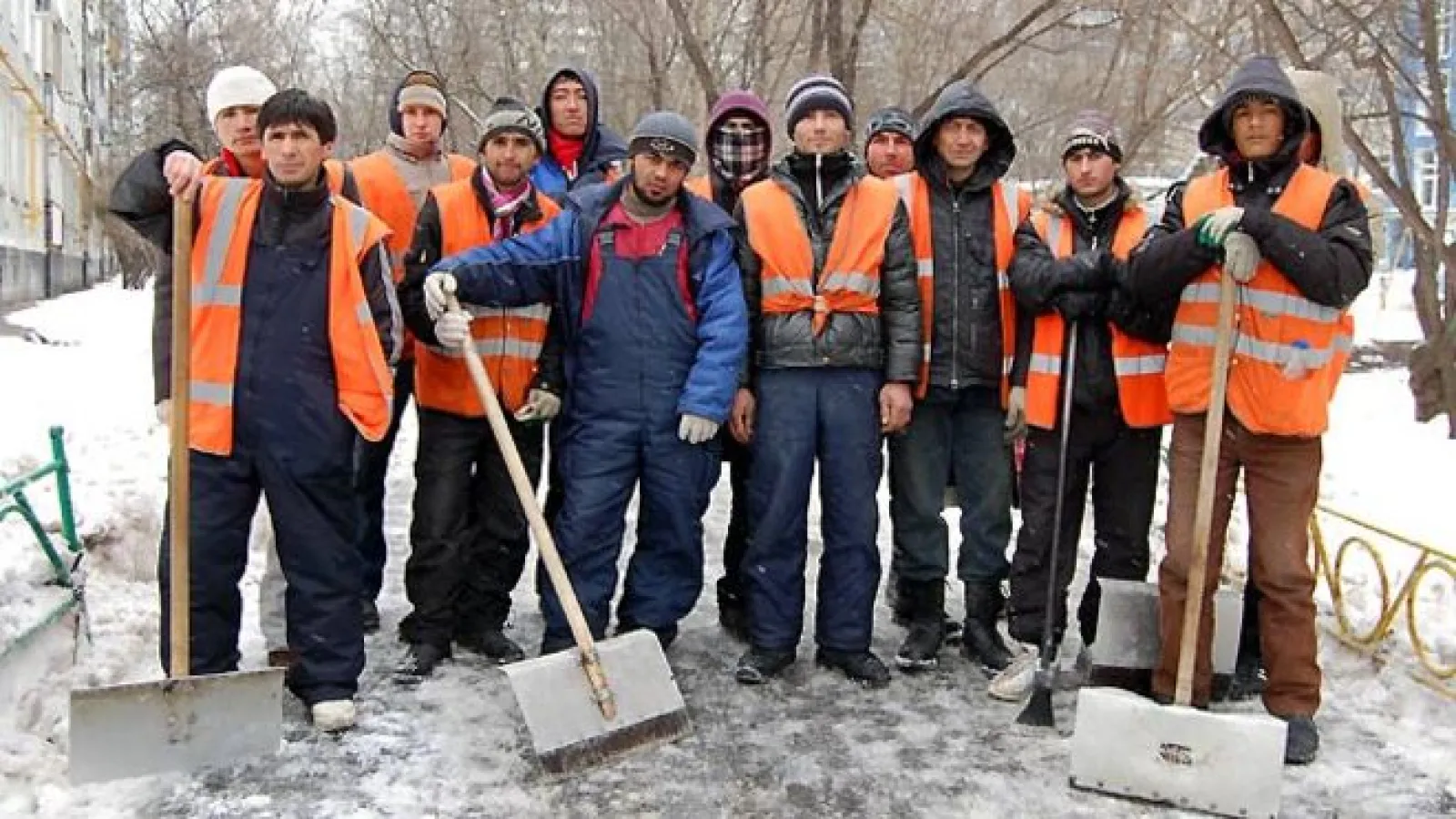 A large group of men standing in rows looking at the camera on a snow street with shovels wearing bright orange vests