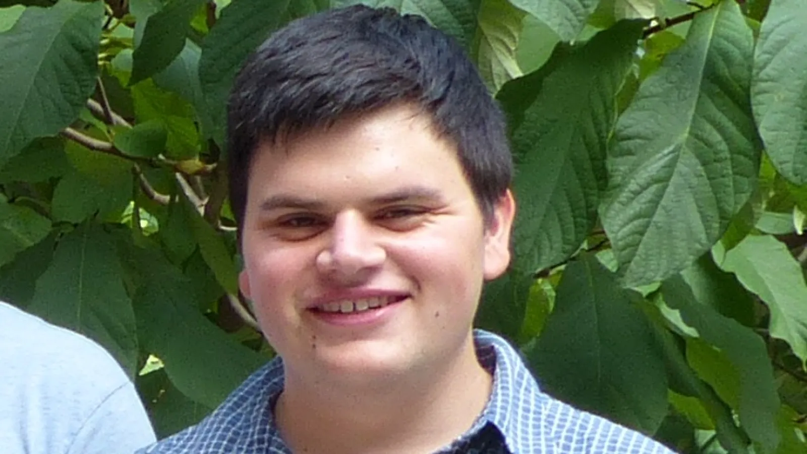 Close up of man's face looking directly at the camera, smiling, with green leaves in the background