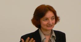 Woman with shorter red hair standing in front of a white background talking