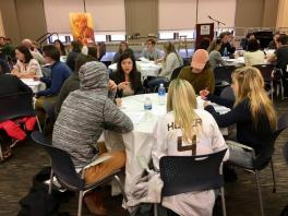 Students sitting around a round table listening to a woman speaking