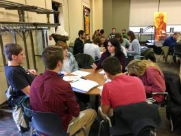 Students sitting around a round table listening to a woman speak