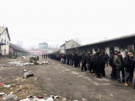 Long line of men standing front-to-back along a long building wearing heavy coats