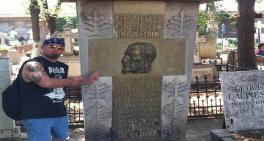 Man wearing sunglasses and a bandana pointing at the large stone tombstone of George Cosbuc