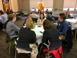 Four young students sitting at a round table listening to a man and woman speak