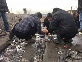 3 men huddles close to the ground, which is covered in garbage, cupping water in their hands coming out of a small pipe