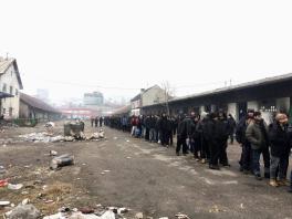 Lunch Line at the Barracks, Photo by Kathryn Metz Long line of men standing front-to-back along a long building wearing heavy coats