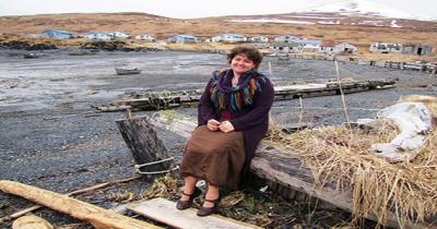 Daria in Akhiok Woman sitting on a piece of timber with a village and mountains far away in background