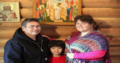 Man, young boy, and woman standing close together in cabin with icons hanging on wall