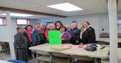 Group of Elders with the Alutiiq Abewenga Group of women standing around a table holding a sign in Alutiiq