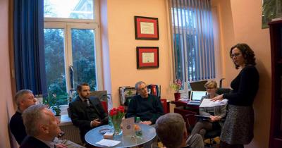 Group of men sitting around a table listen to woman speak who is standing nearby 