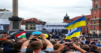 Crowded square with stage in distance with screening showing the president speaking