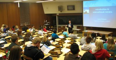 Students sitting at desks in a large auditorium looking at man standing in front speaking by projector screen