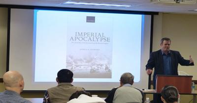 Man standing next to a large projector screen that is showing a bookcover and words Imperial Apocalypse