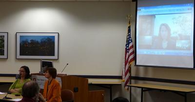 Two women sitting at a table on the left with a large projector screen on the right showing a woman's face speaking