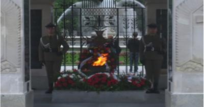 Two soldiers in uniform standing guard on either side on a stone monument with a flame and surrounded by bouquets