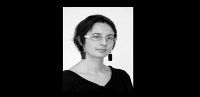 A black and white image of a woman with short hair, glasses, and large rectangular earrings look at the camera