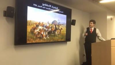 Man standing at a podium talking, looking at a video screen showing presentation visuals