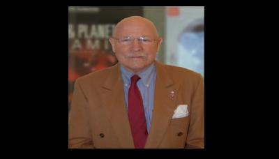 A man wearing a brown suit coat and wearing glasses looking a tthe camera with large, blurred posters in the background