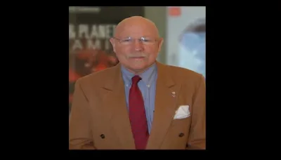 A man wearing a brown suit coat and wearing glasses looking a tthe camera with large, blurred posters in the background