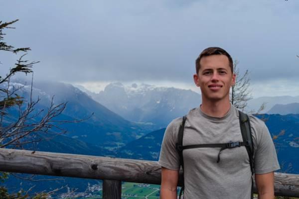 Yasha Shatalov stands wearing a backpack in front of a green mountain landscape