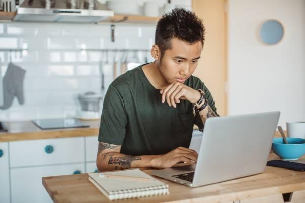 A young man with spiky black hair and tatoos wearing a dark green t-shirt works at a laptop