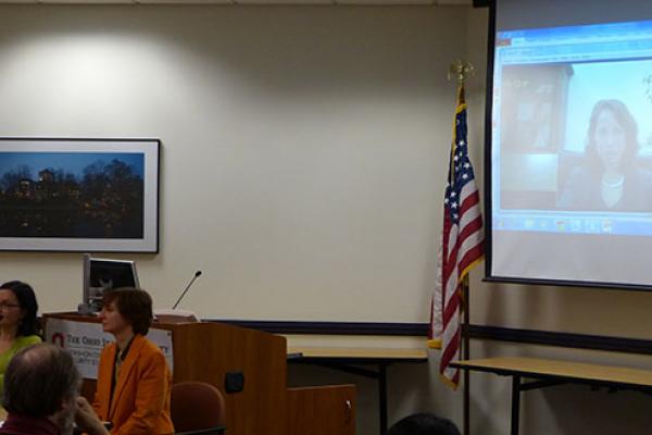 Two women sitting at a table on the left with a large projector screen on the right showing a woman's face speaking