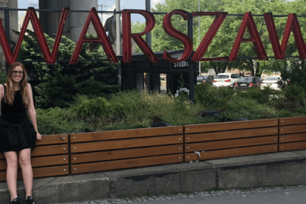 A girl with long hair and glasses standing by a large sign spelling out Warszawa