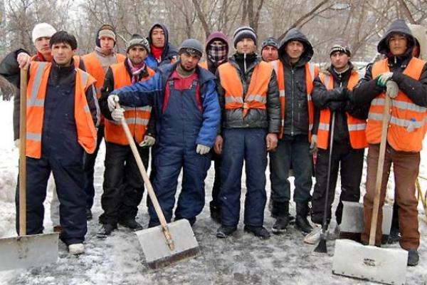 A large group of men standing in rows looking at the camera on a snow street with shovels wearing bright orange vests