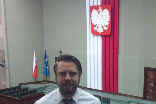 Peter Tunkis in the Sejm A man standing overlooking a gallery with a flag hanging in background.