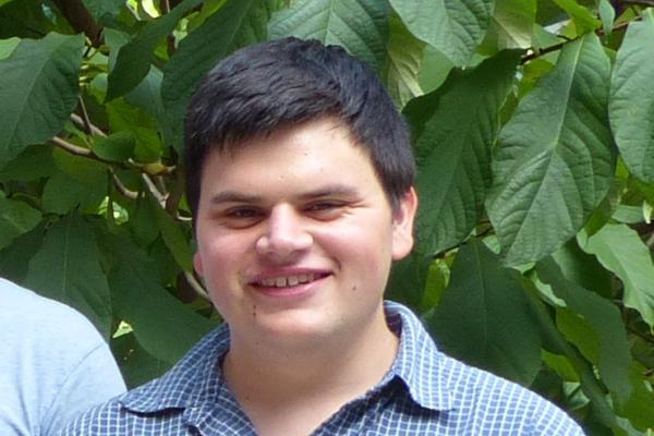 Close up of man's face looking directly at the camera, smiling, with green leaves in the background