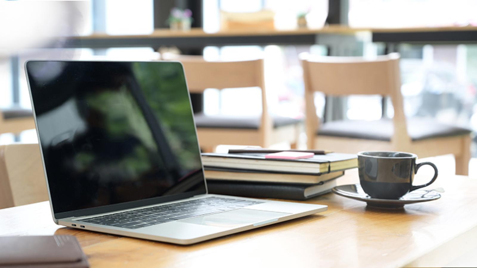 Open laptop sitting on a wooden desk with a stack of books and teacup placed to the side of it.