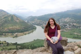 Girl standing on a high hill facing camera with a winding river, towns, and mountains in the background